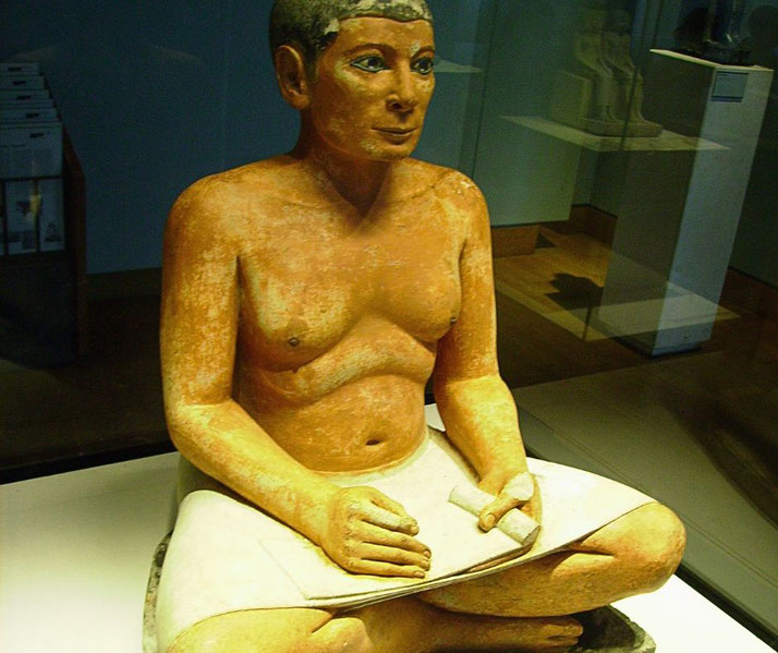 A painted statue of an ancient Egyptian scribe sitting cross-legged, holding a papyrus scroll and stylus, displayed in a museum exhibit. The figure has detailed facial features and a lifelike expression.