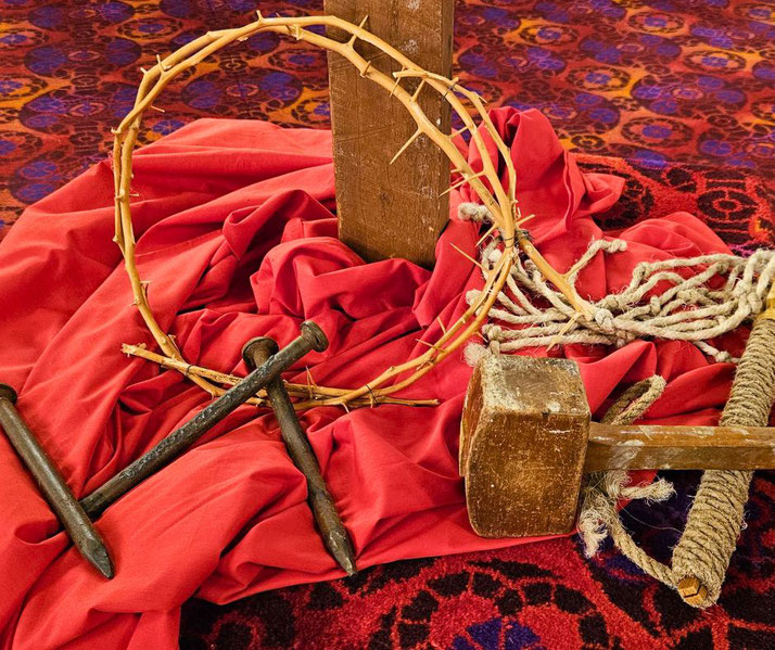 A symbolic display of the crucifixion of Jesus, featuring a crown of thorns, large nails, a wooden mallet, a whip, and a cross, all placed on red fabric with a patterned carpet.