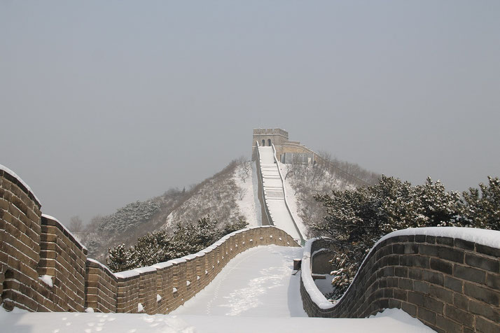 The Great Wall of China covered in snow, stretching over a winter landscape. A watchtower stands in the distance, while footprints mark the untouched white path along the wall.