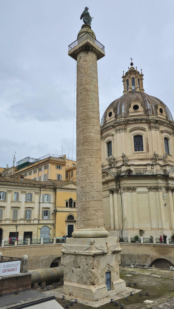 A tall Roman column with spiral reliefs stands in a plaza, topped by a statue and surrounded by historic buildings, including a domed church in the background.