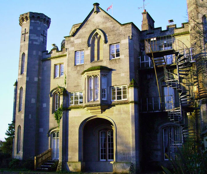 A Gothic Revival-style building with pointed arches, a turret featuring a cross-shaped window, and a spiral staircase. The structure is adorned with ivy and lit by sunlight.