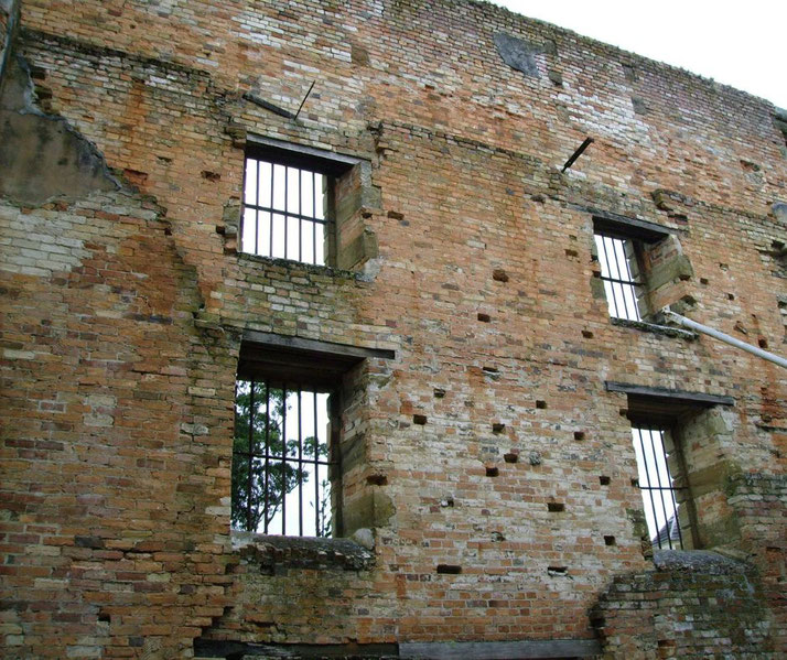 A crumbling brick wall with barred windows stands weathered and damaged, showing missing bricks and decay.