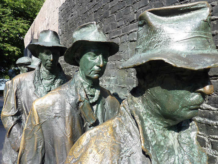 Bronze statues of men in hats stand in a line against a brick wall, depicting a breadline scene from the Great Depression.