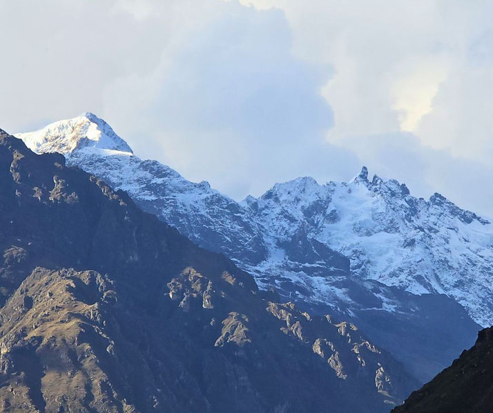A stunning view of snow-capped mountains with rugged, shadowed slopes in the foreground. The peaks rise against a cloudy sky, creating a dramatic contrast between the icy summits and dark terrain.