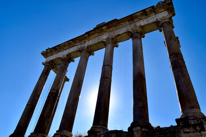 The towering ruins of an ancient Roman temple, with Corinthian columns silhouetted against a bright blue sky.