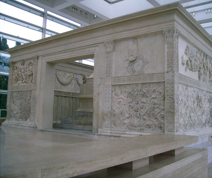 Side view of a Roman marble altar with detailed relief carvings, floral patterns, and mythological figures inside a modern glass building.