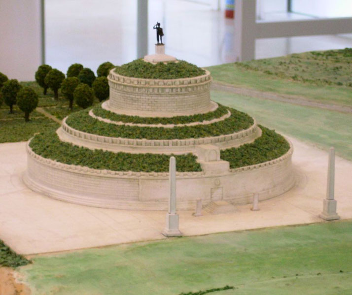 Scale model of a circular, tiered Roman mausoleum with greenery and a statue on top, surrounded by pathways and small obelisks.