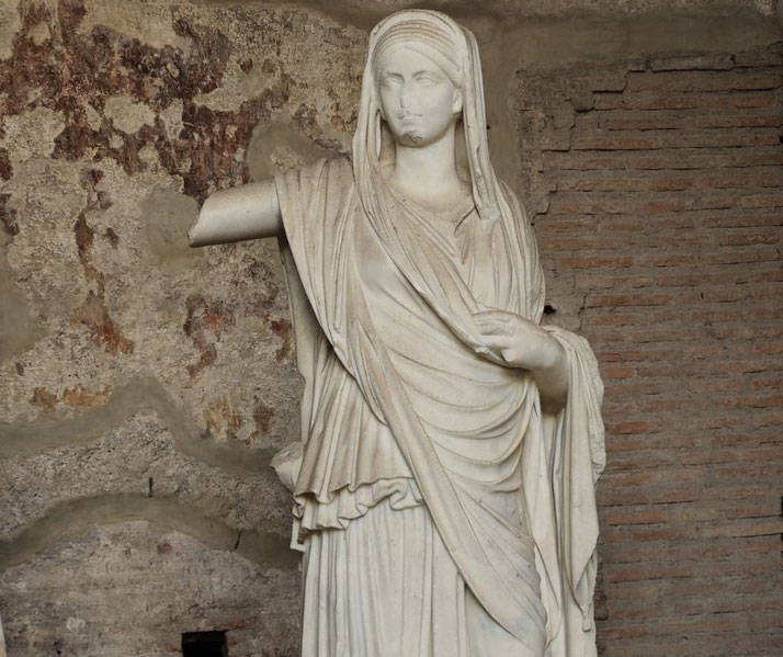 Ancient marble statue of a veiled woman with one missing arm, draped in flowing robes, set against a weathered stone and brick wall.