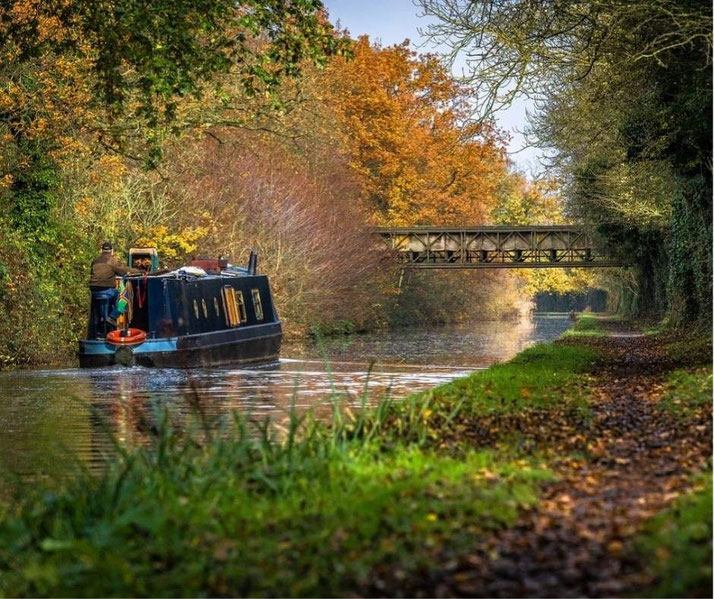 peaceful canal surrounded by autumn foliage. A metal bridge spans the water, and a towpath lined with grass and fallen leaves follows the canal’s curve into the distance.