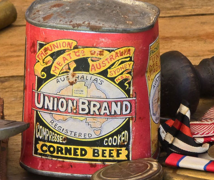 A vintage red tin of Union Brand compressed cooked corned beef from Australia sits on a wooden surface near medals and artifacts.