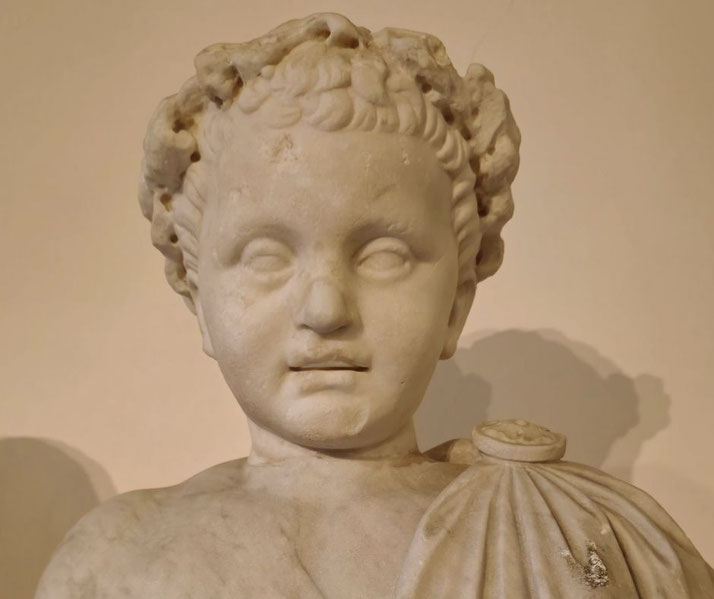 Marble bust of a young child with curly hair and chubby cheeks, wearing a draped garment fastened with a round brooch on the shoulder.