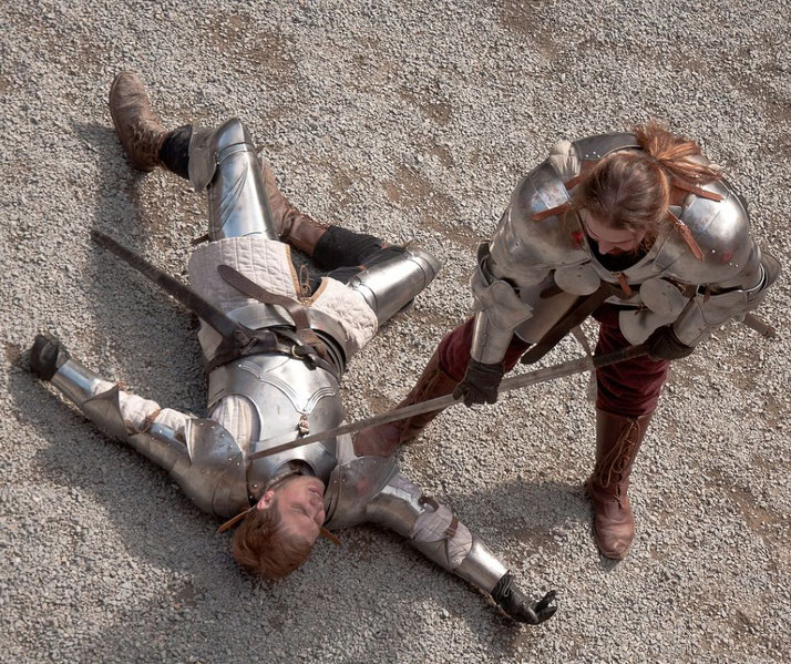 Two armored knights engage in a duel. One lies on the ground defeated, while the other stands over them, sword pointed at their chest.