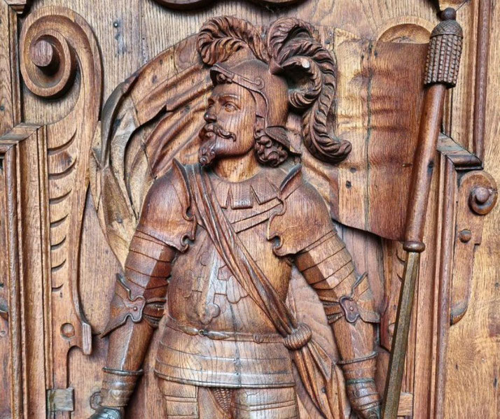 An intricately carved wooden figure of a medieval knight, adorned in armor with a plumed helmet, holding a staff or banner, surrounded by ornate decorative details.