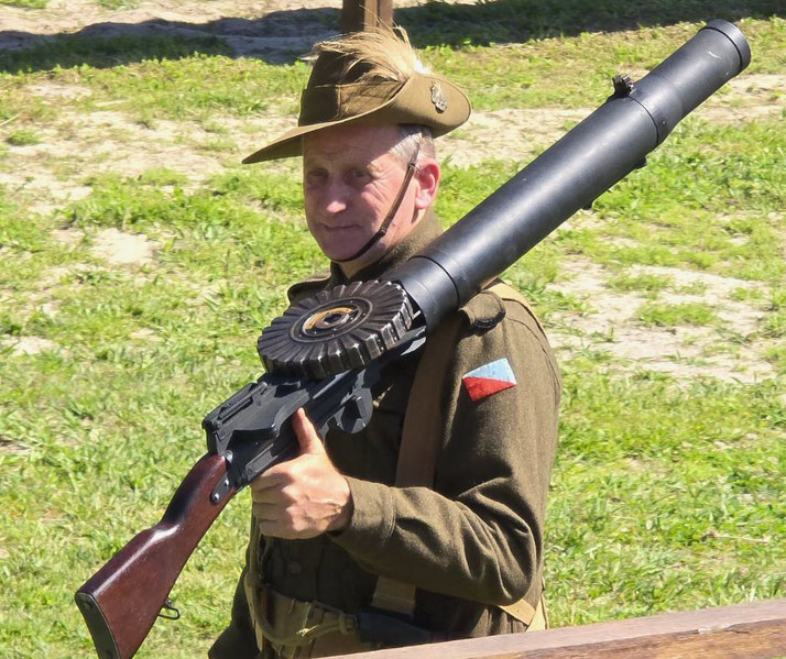 A man in vintage military uniform carries a Lewis gun with a distinctive round drum magazine and wooden stock over his shoulder.