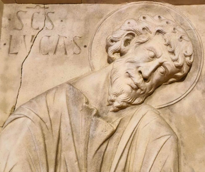 A carved stone relief depicting Saint Luke ("SCS LVCAS"), with a halo around his head. The figure has a solemn expression, curly hair, and a beard, with visible cracks in the aged stone surface.