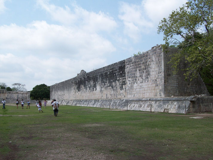 Great Ball Court of Chichen Itza, a large ancient Mayan ball court with tall stone walls. Visitors walk along the grassy field, where the Mesoamerican ball game was once played in ritual contests.