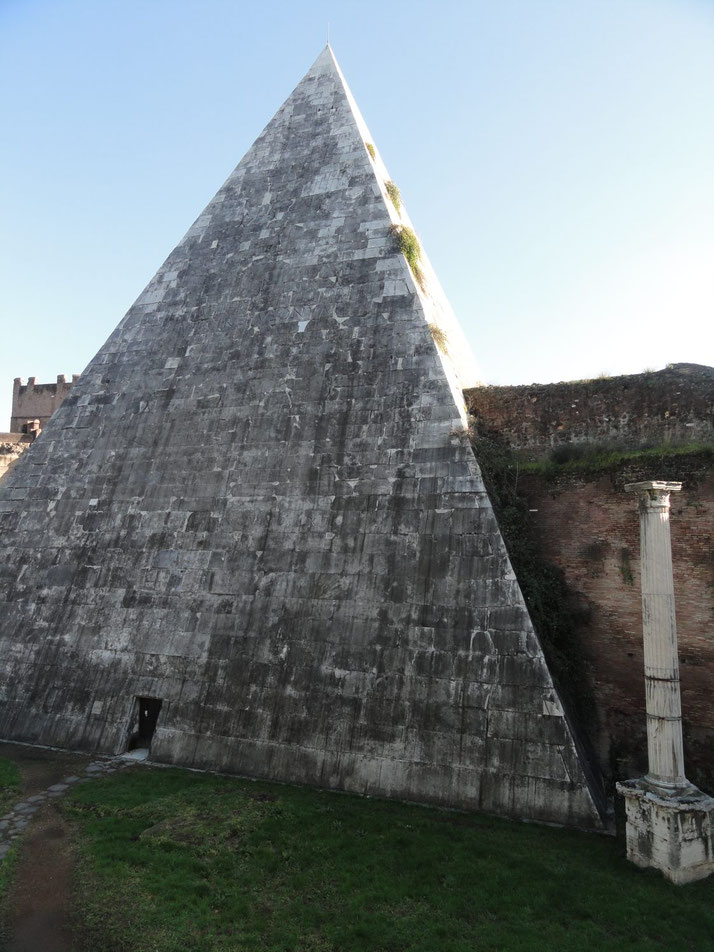 Close-up of a tall stone pyramid with a small entrance at the base, next to a single ancient column and a brick wall under a bright sky.