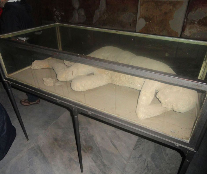 Plaster cast of a victim from Pompeii displayed in a glass case, showing the preserved pose at the time of the volcanic eruption.