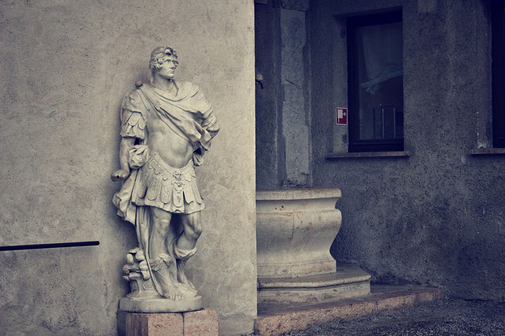 Marble statue of a Roman figure in armor stands against a textured wall near a window and stone basin.