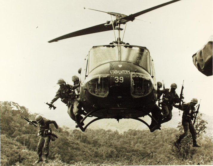 Soldiers jump from a hovering military helicopter labeled “Headhunters 39” into a jungle clearing, rifles ready for action.
