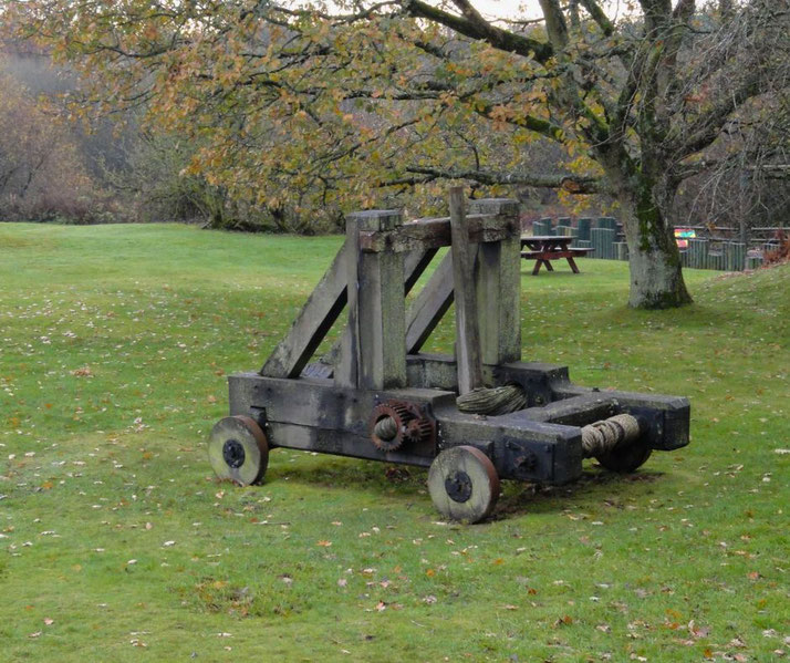 Old wooden catapult sits on grass in a park-like setting, surrounded by trees and picnic tables in the background.