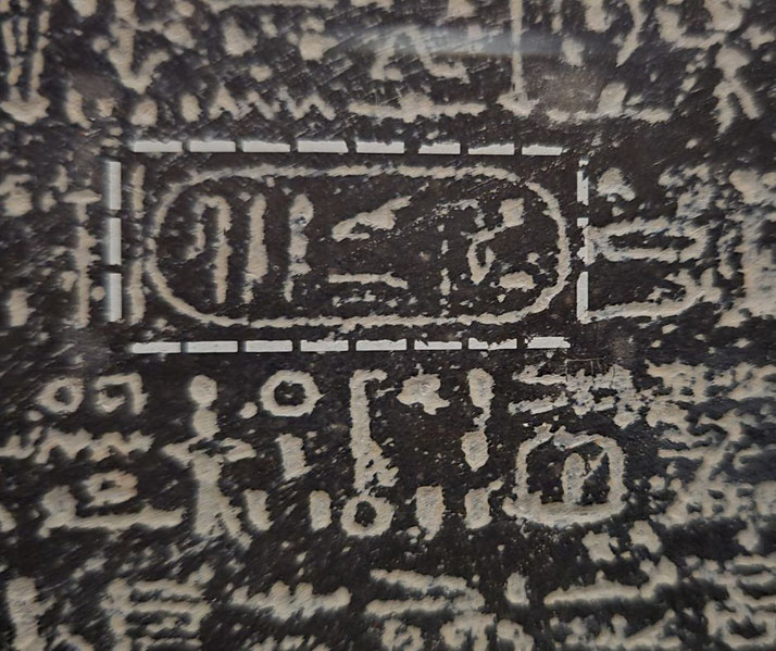 A close-up of ancient Egyptian hieroglyphs on a dark stone surface, featuring a cartouche enclosing a royal name. The inscription appears weathered but retains detailed carvings.