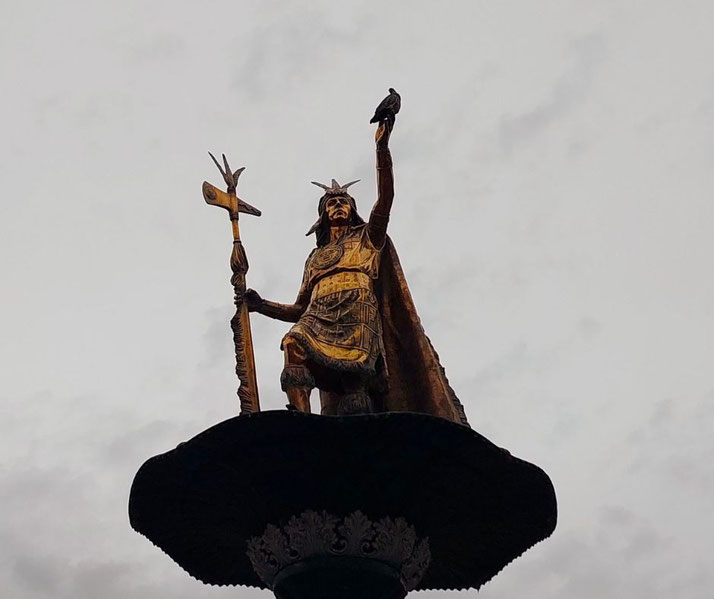A golden statue of Inca ruler Pachacuti stands atop a fountain in Cusco, Peru. He holds a staff and raises his hand, where a pigeon is perched, against a cloudy sky.