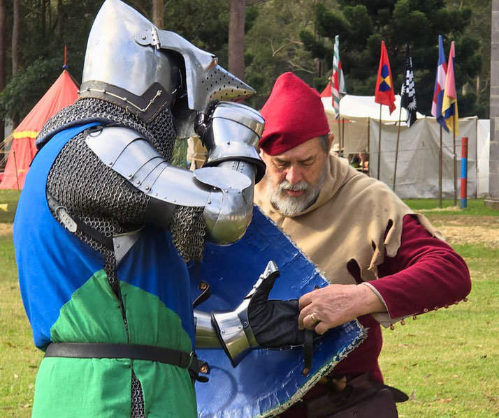 A knight in blue and green armor being assisted by an older man in medieval attire. The knight wears chainmail and a steel helmet, while the man helps adjust a gauntlet.