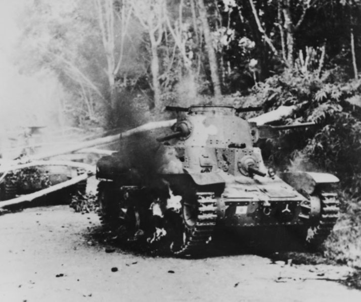 A damaged tank burns on a jungle road, with smoke rising from its side after being hit in combat.