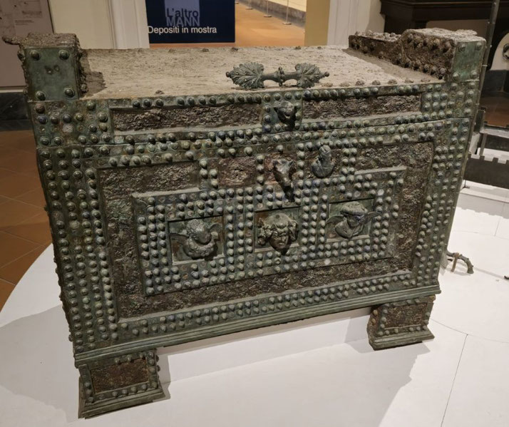 Ancient bronze chest adorned with rows of decorative studs and relief faces, featuring a weathered surface and ornate detailing.