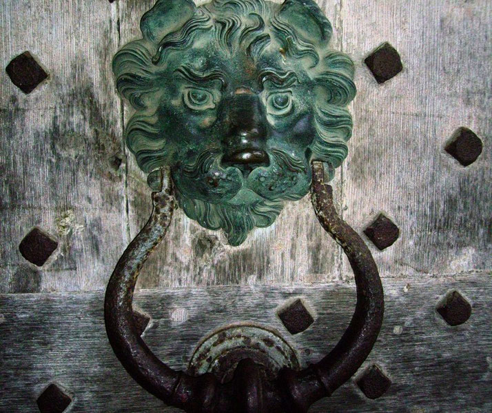 A weathered metal lion-shaped door knocker with a patina finish, mounted on an old wooden door with dark decorative studs, exuding an antique and rustic charm.