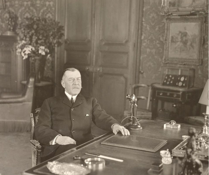 Black and white photograph of a middle aged man in a suit sitting at a desk with an early telephone, formal office interior, patterned walls, framed artwork, and period furniture.
