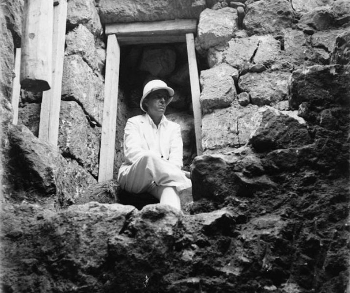 A person in light clothing and a pith helmet sits in a stone doorway surrounded by rough masonry, likely at an archaeological site.