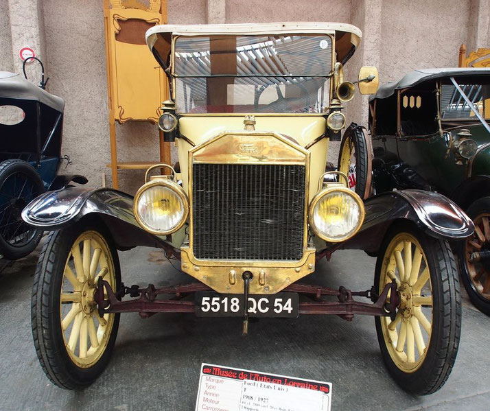 A yellow and black 1927 Ford Model T is on display in a museum, viewed from the front with brass detailing and large round headlights.