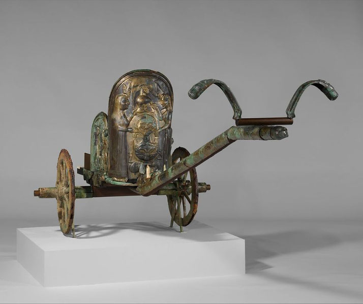 An ancient bronze chariot with intricate relief decorations, including human figures and a central face motif.