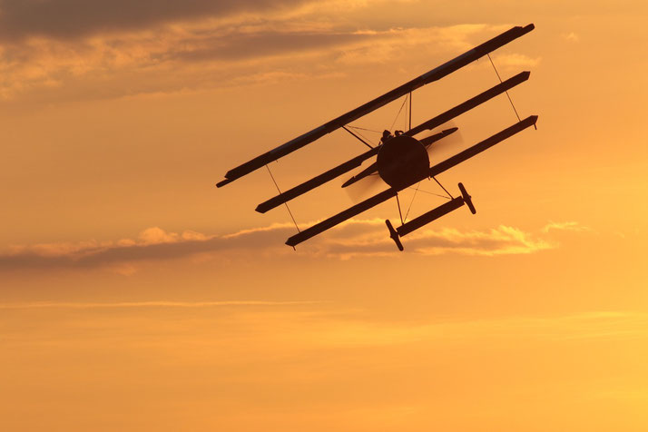 Silhouette of a World War I-era triplane flying against a golden sunset sky, with its three wings and open cockpit clearly visible.