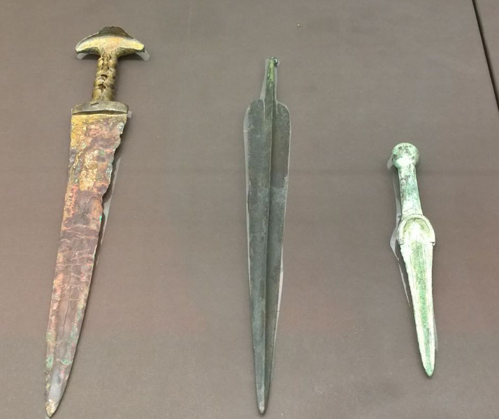 Three ancient bronze daggers with distinct blade shapes and patinas displayed on a flat surface.