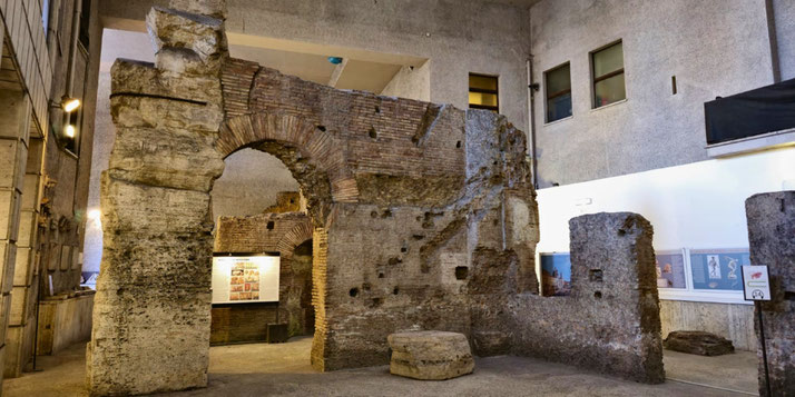 Ancient Roman brick and stone ruins displayed indoors, featuring an arched doorway and partial walls, surrounded by modern museum lighting and informational panels.