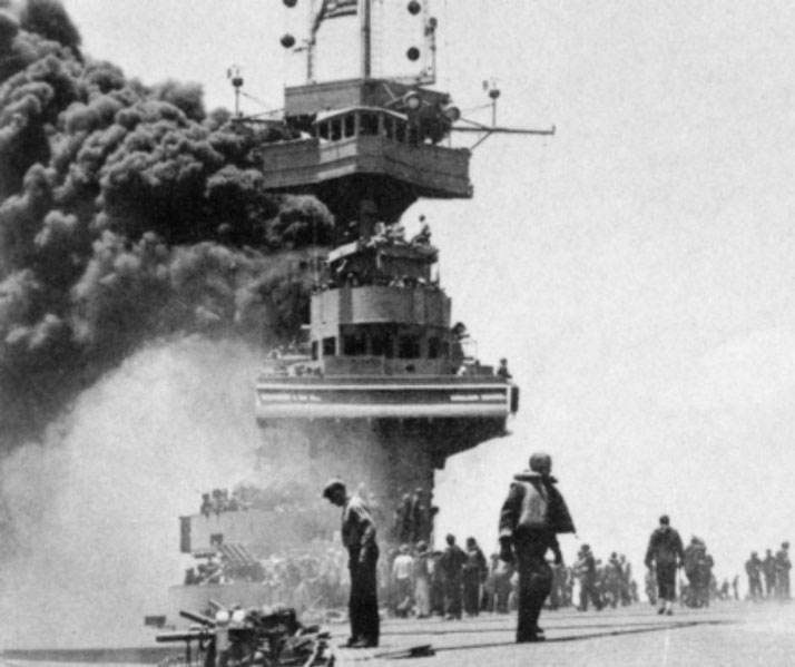Sailors gather on the deck of an aircraft carrier as thick smoke rises from an onboard fire during battle.