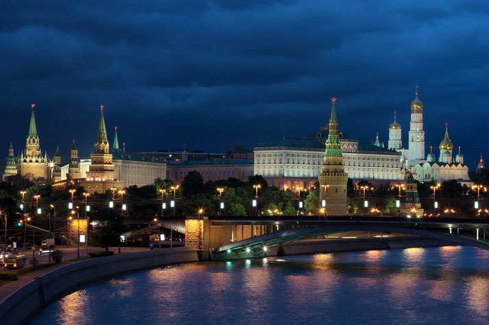 Night view of the Kremlin complex in Moscow with its illuminated towers, domes, and buildings reflected in the river.