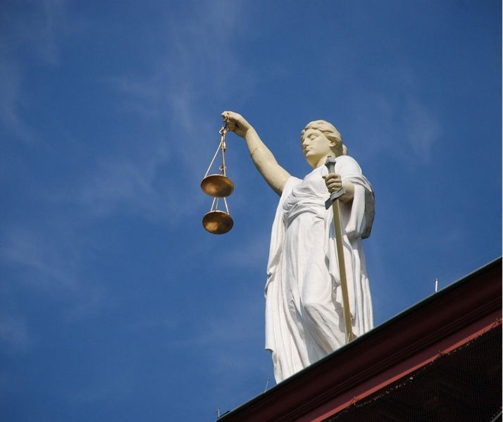 A white statue of Lady Justice stands atop a building, holding balanced scales in one hand and a sword in the other against a clear blue sky.