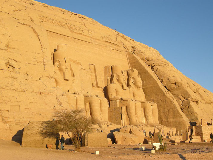 Wide view of Abu Simbel temple carved into a sandstone cliff, featuring colossal seated statues of Ramesses II with people nearby for scale.