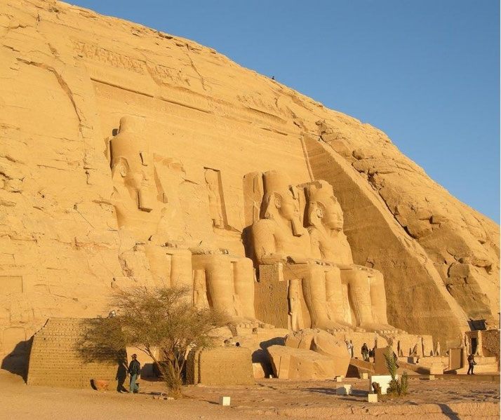 Wide view of Abu Simbel temple carved into a sandstone cliff, featuring colossal seated statues of Ramesses II with people nearby for scale.