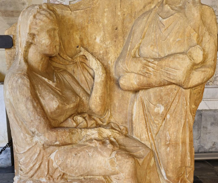 Ancient marble relief of two women in draped robes, one seated with a hand raised, the other standing with arms crossed, both with worn facial features.