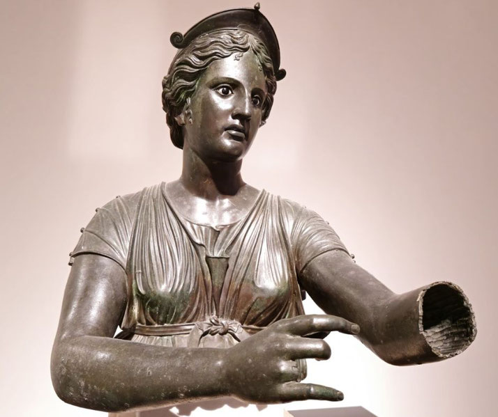 Bronze statue of a woman wearing a crested helmet and draped dress, with one arm missing and the other extended, fingers delicately positioned.