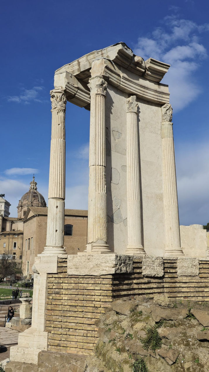 Ruins of an ancient Roman temple with four tall Corinthian columns and a partial entablature, standing on a layered brick and stone base under a clear blue sky.