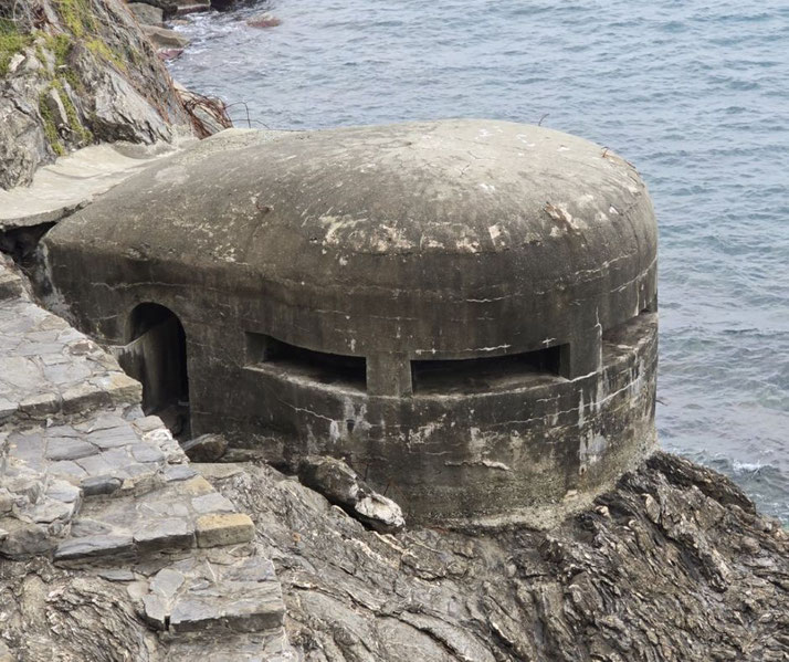 A concrete World War II-era bunker sits on rocky cliffs above the sea, partially embedded in stone with narrow viewing slits.