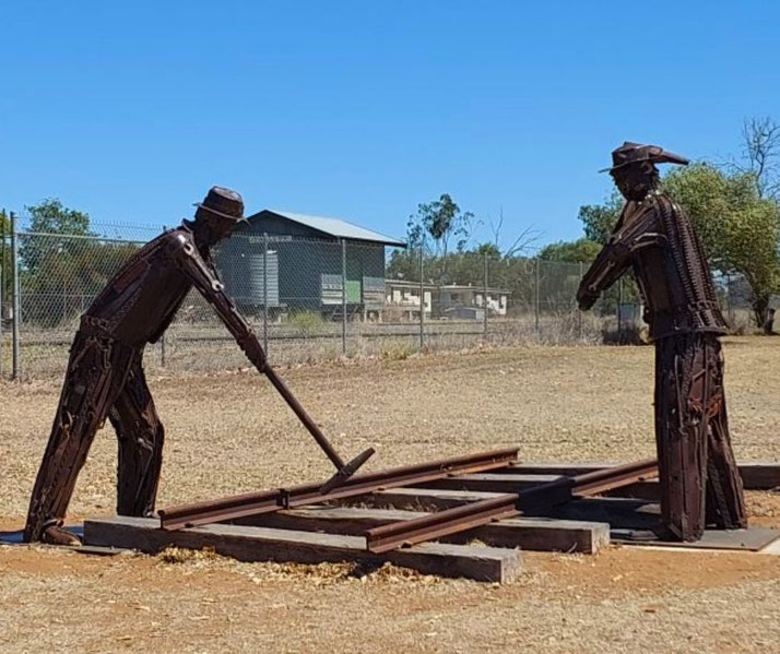 A metal sculpture depicts two railway workers laying tracks, symbolizing Australia's railway heritage. Made from scrap metal, it stands in a rural setting, honoring the laborers who built the railways.