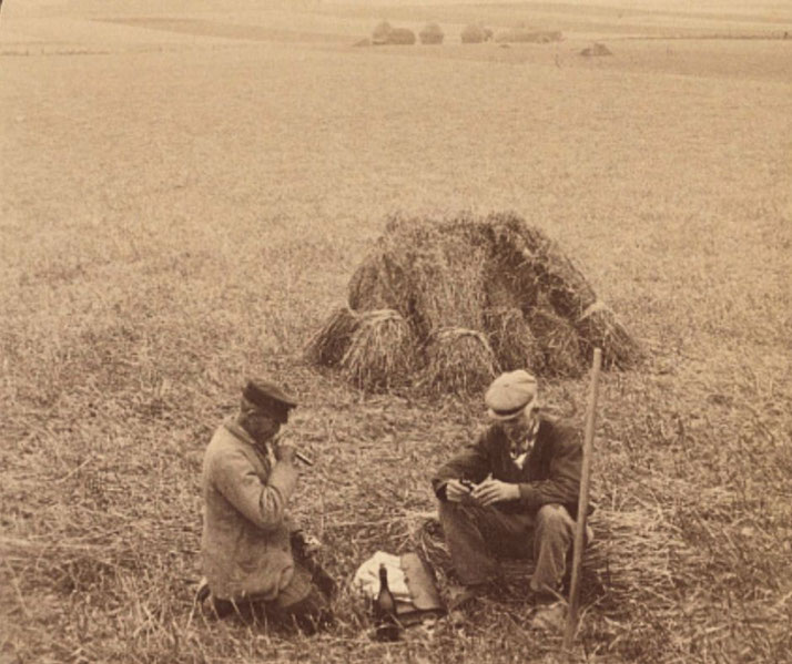 A vintage sepia-toned photograph of two farmworkers taking a break in a field. They sit near a pile of harvested crops, one smoking a pipe and the other rolling a cigarette, with tools and a bottle beside them.
