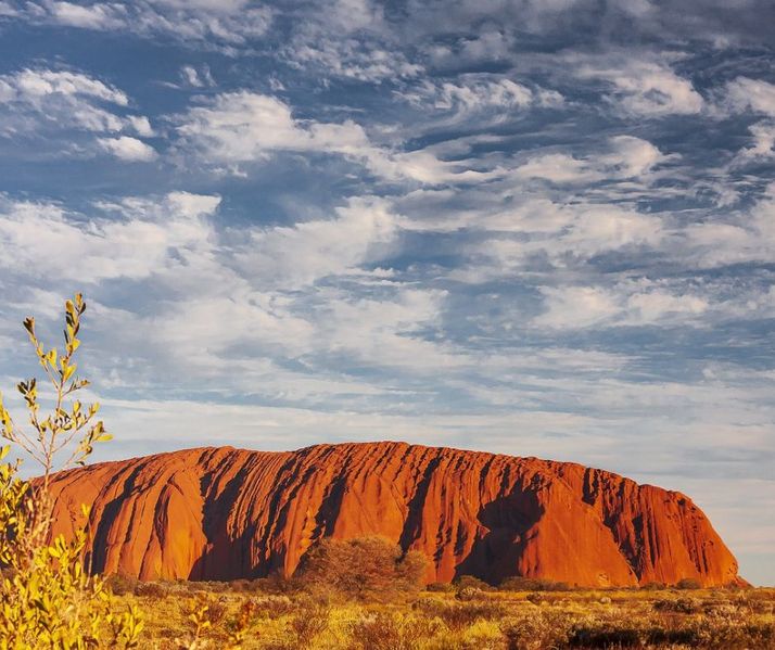 Uluru, Australia's iconic red sandstone monolith, bathed in warm sunlight. The rugged formation contrasts with a sky filled with wispy clouds, creating a stunning desert landscape.